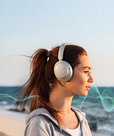 Mujer con auriculares blancos Over-Ear en la playa, el viento sopla entre su cabello.