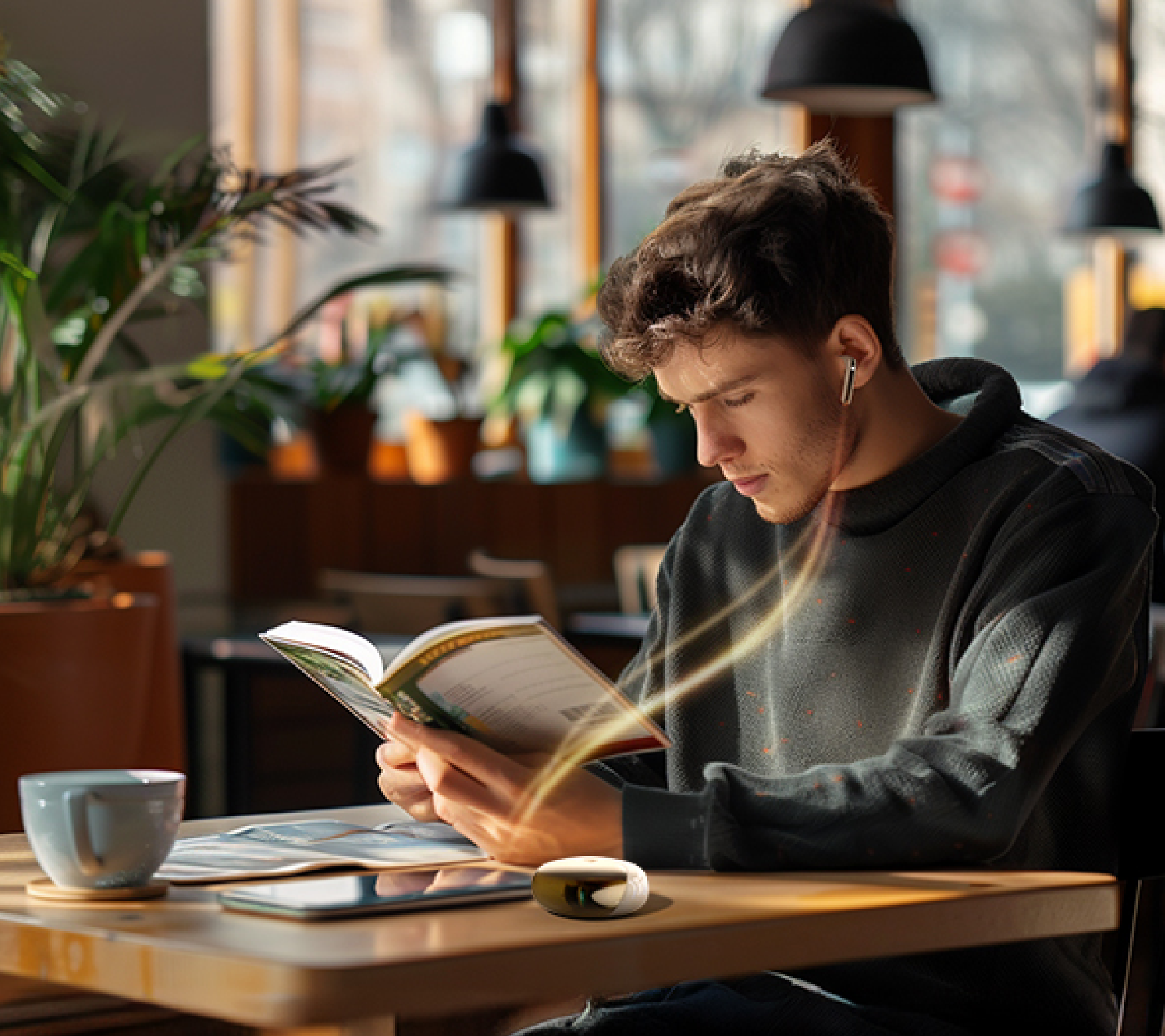 Joven leyendo un libro en una cafetería con auriculares inalámbricos y una taza de café sobre la mesa de madera.