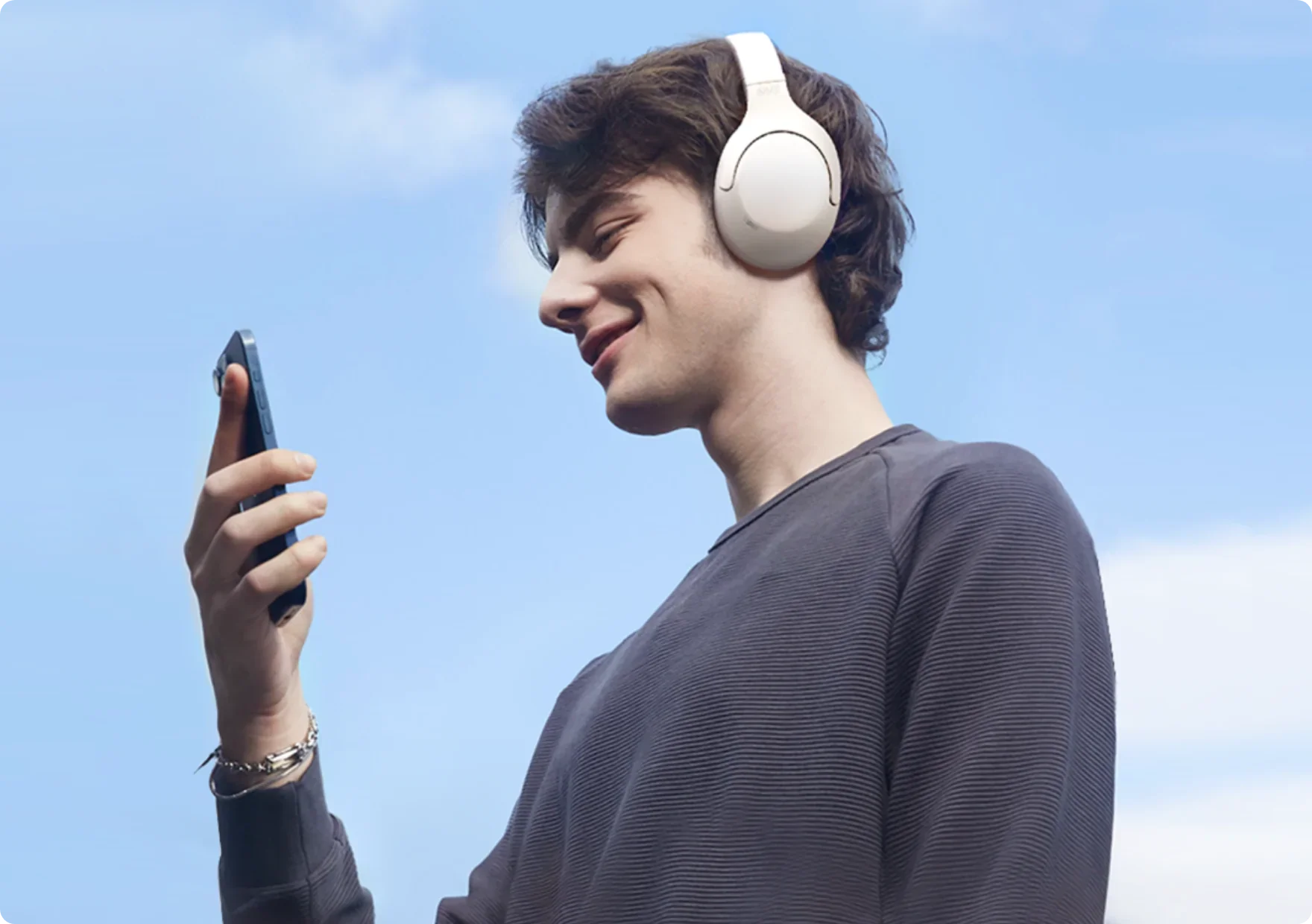 Joven con auriculares blancos y smartphone bajo un cielo azul.
