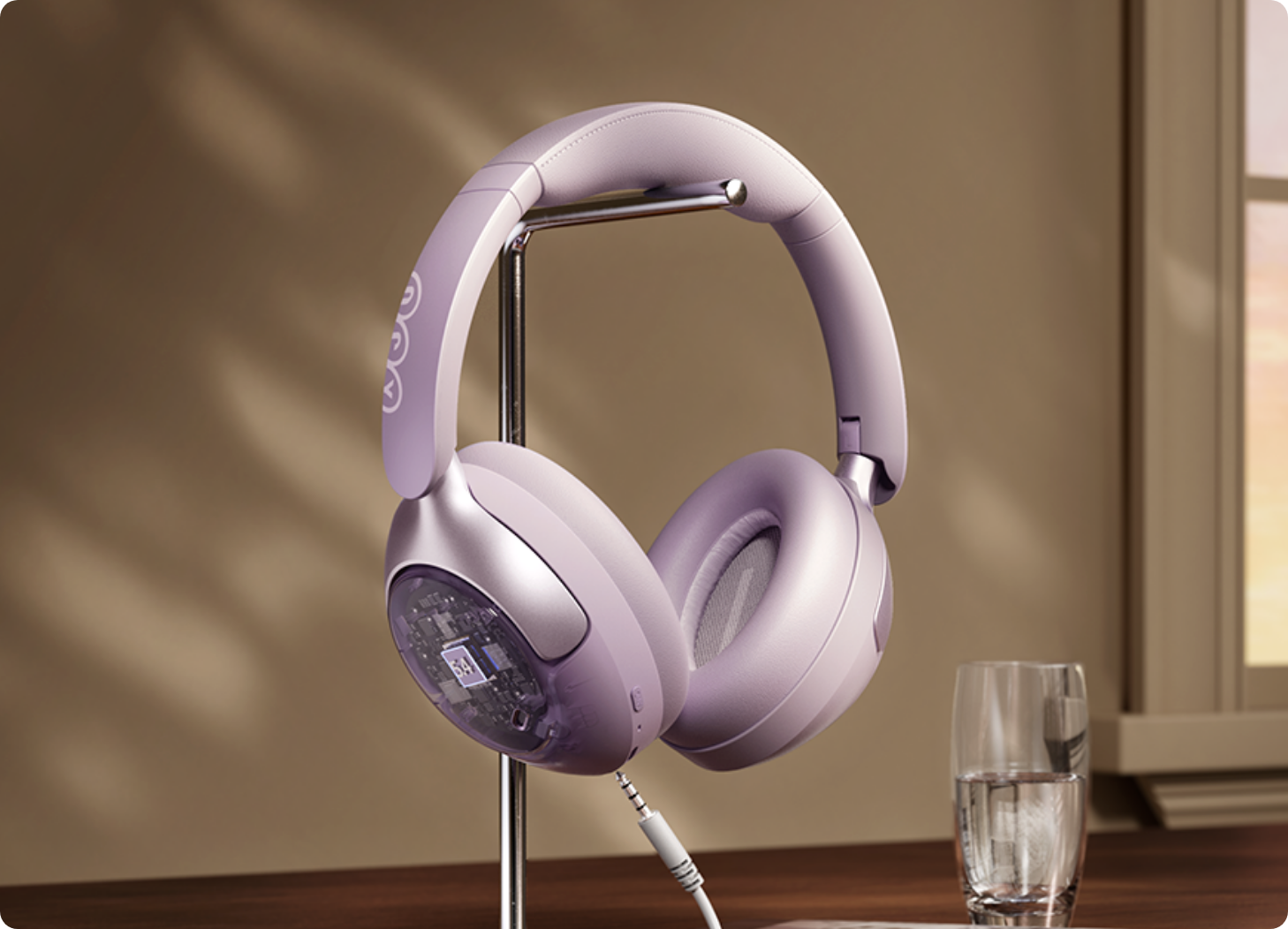 Auriculares over-ear color lavanda con carcasa transparente en soporte, cable conectado, junto a un vaso de agua en la ventana.